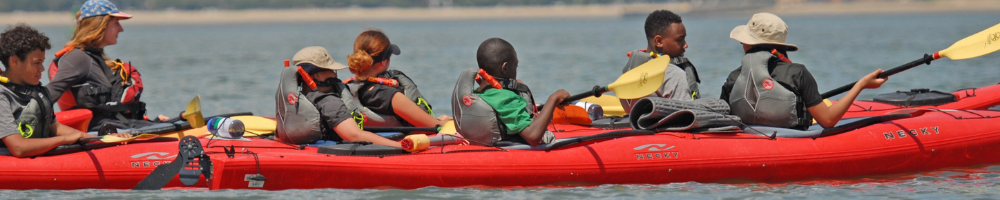 The image shows a group of people kayaking on a lake. They are in a long, red kayak and appear to be enjoying a sunny day. The water is calm, and the background shows a distant shoreline. Everyone is wearing life vests.